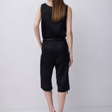 A woman is shown from the back wearing a black sleeveless top and matching black cropped pants with a thin dark belt, paired with black strappy heels, against a light gray studio background.