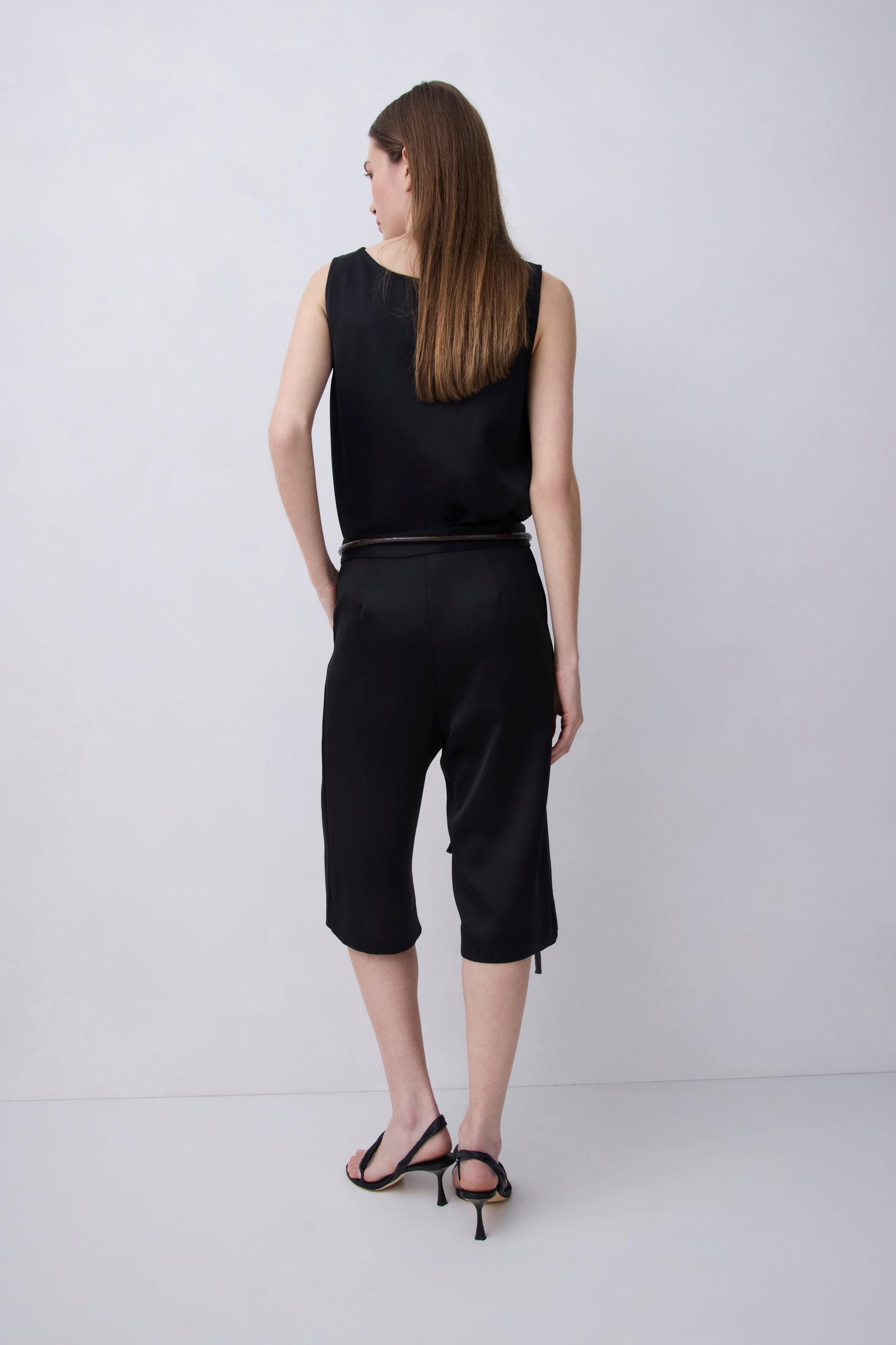 A woman is shown from the back wearing a black sleeveless top and matching black cropped pants with a thin dark belt, paired with black strappy heels, against a light gray studio background.