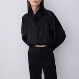 A model wearing a black cropped jacket with a high gathered collar and voluminous sleeves, paired with black cropped trousers, standing against a light gray studio background.