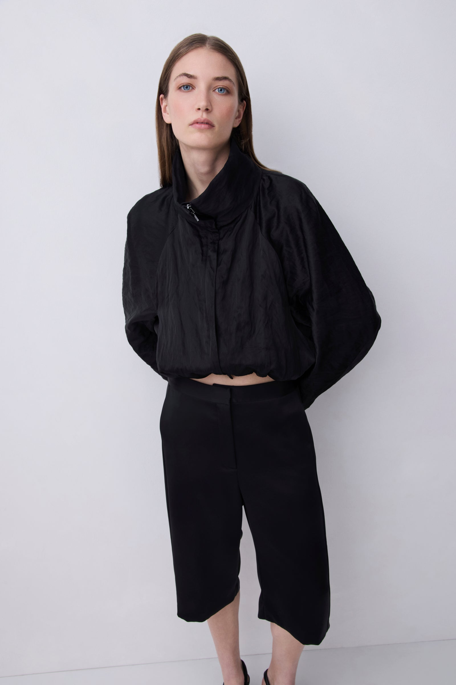 A model wearing a black cropped jacket with a high gathered collar and voluminous sleeves, paired with black cropped trousers, standing against a light gray studio background.