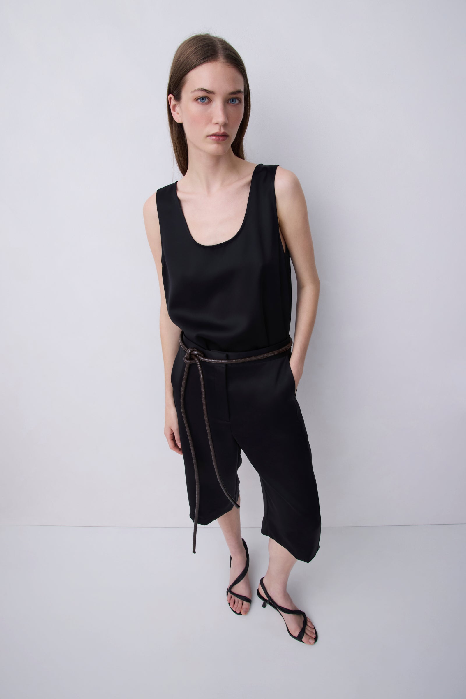 A model wearing a black sleeveless scoop-neck top and matching black cropped pants with a thin brown belt, paired with black strappy sandals, against a light gray studio background.