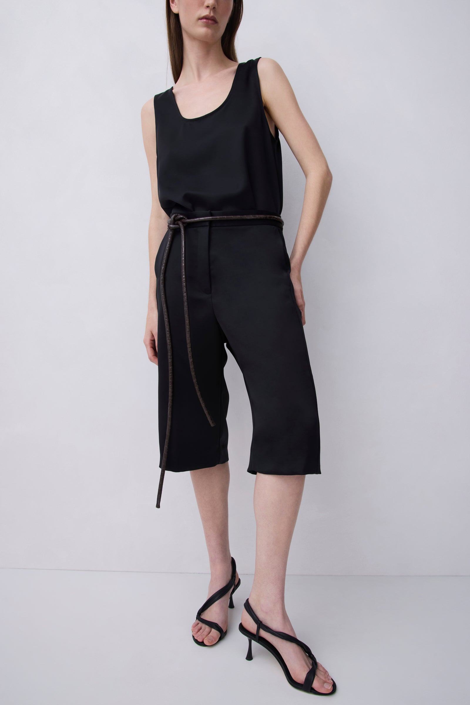 A model wearing a black sleeveless top and matching black cropped pants with a thin dark tie belt, paired with black strappy high-heeled sandals, against a light gray studio background.