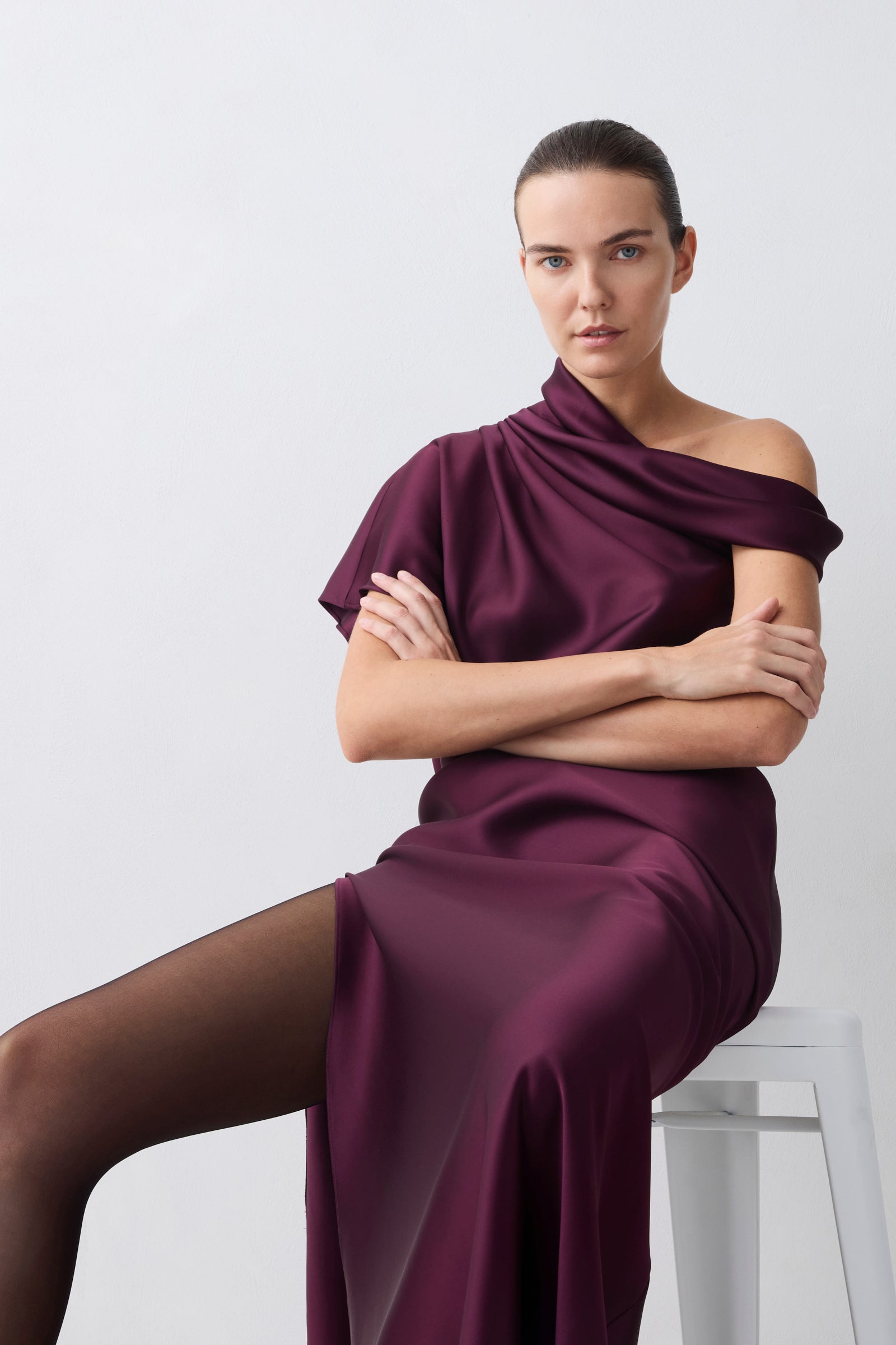 Woman wearing a burgundy off-shoulder dress sitting on a white stool against a light gray background