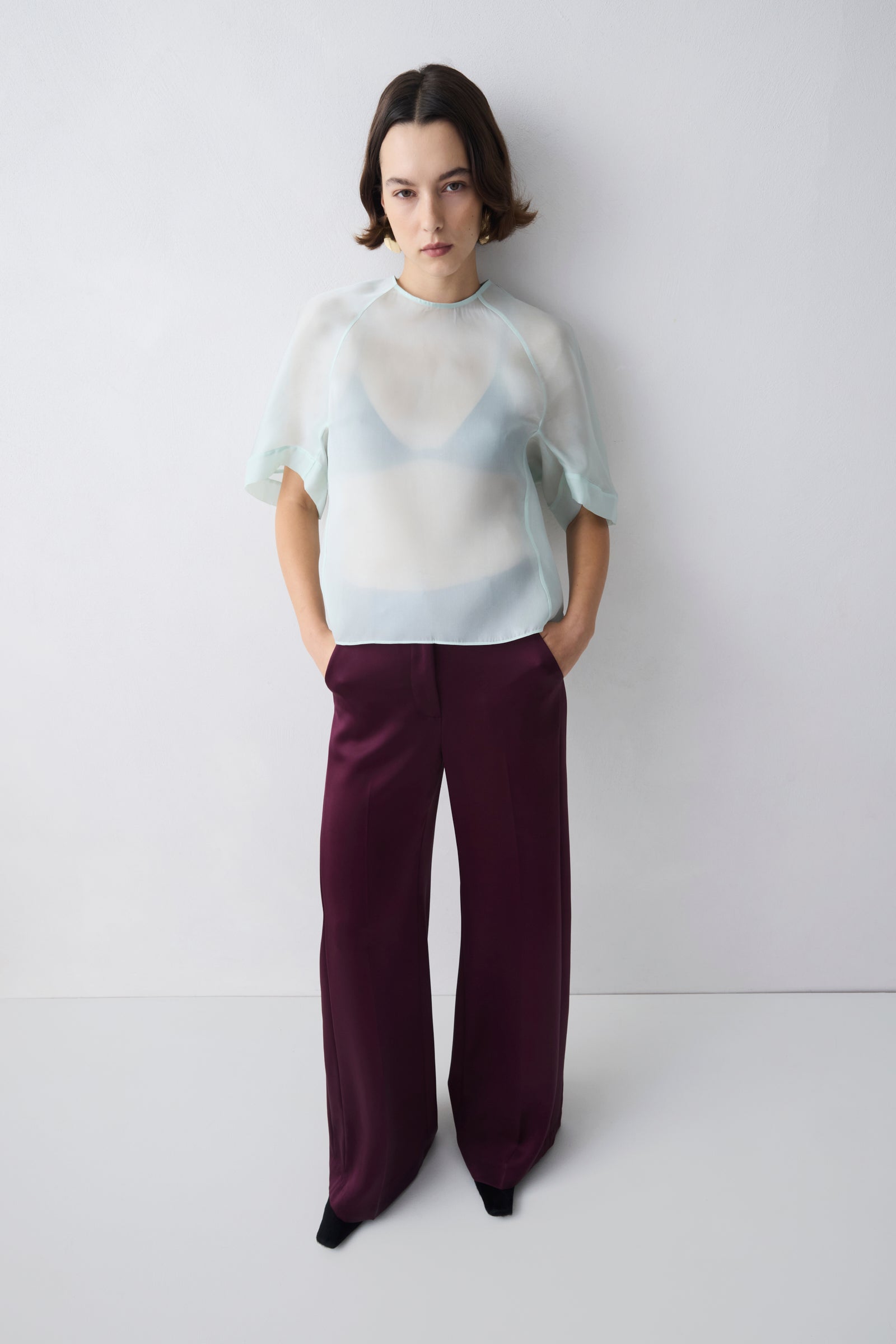 Person wearing a sheer white top and dark purple pants against a white background