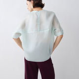 Person wearing a light gray blouse with a subtle pattern and dark purple pants on a white background