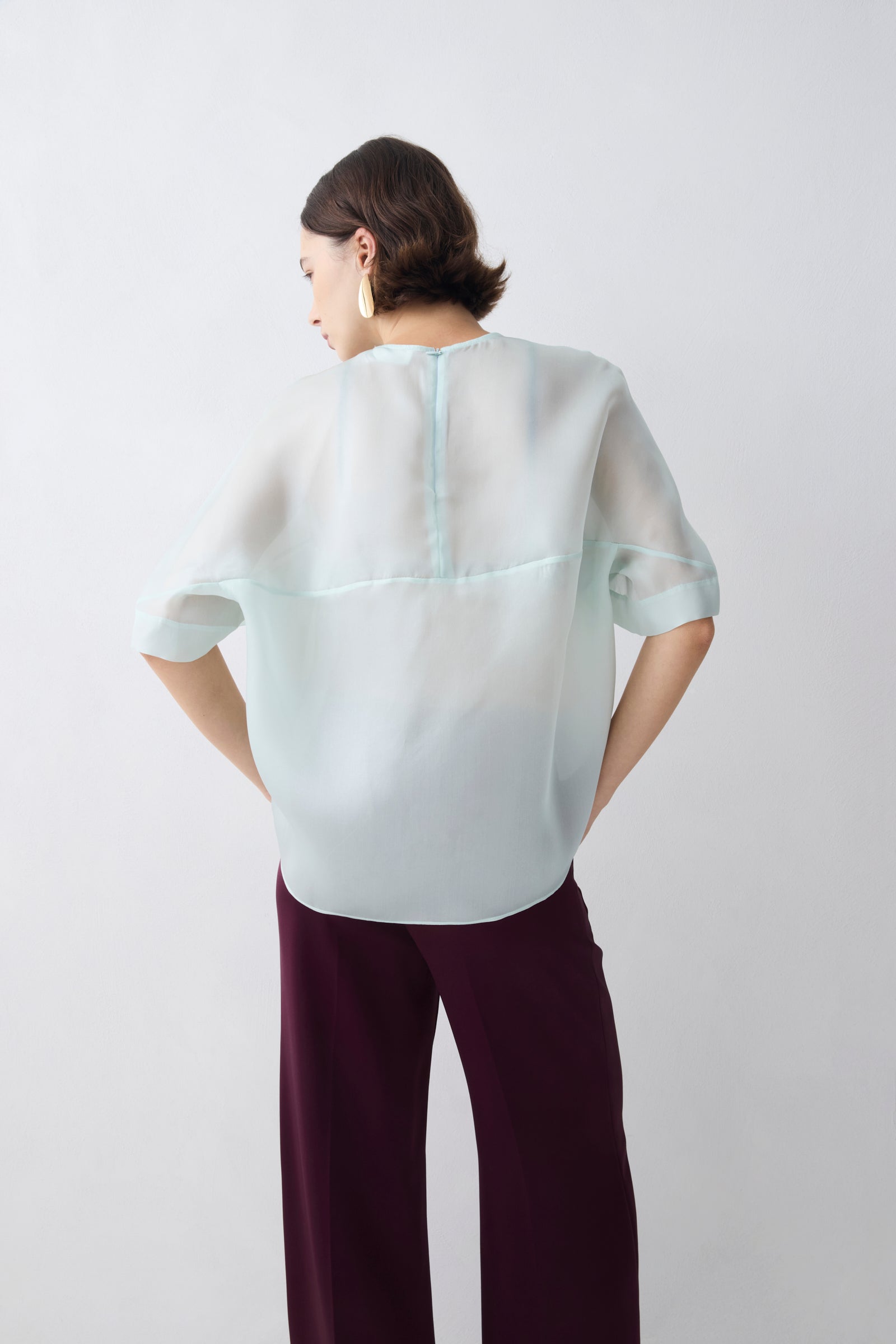 Person wearing a light gray blouse with a subtle pattern and dark purple pants on a white background