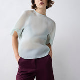 Woman wearing a light-colored blouse and dark pants against a plain background