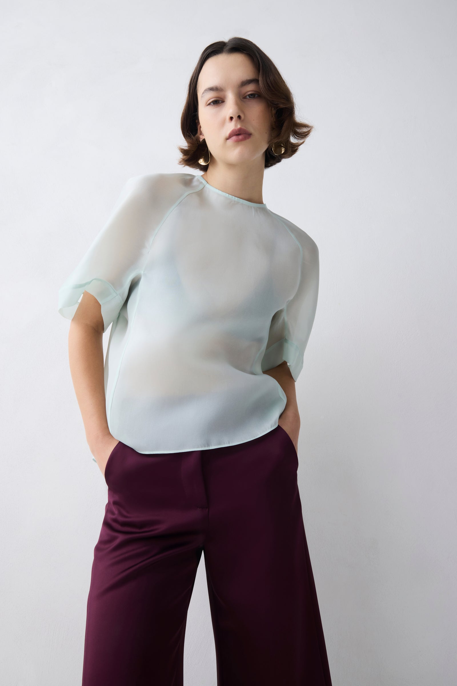 Woman wearing a light-colored blouse and dark pants against a plain background