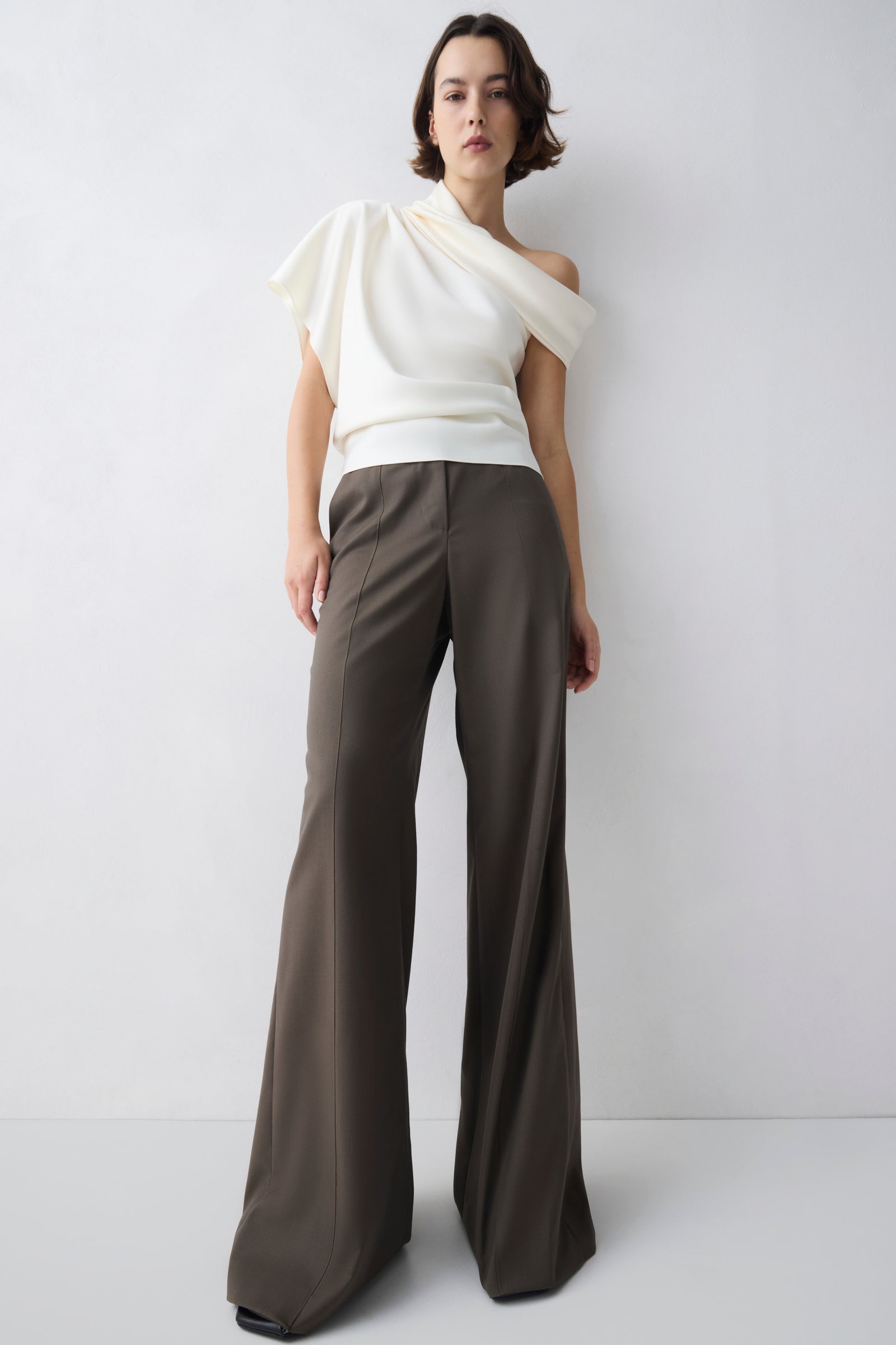 Woman wearing a white top and brown pants against a plain background