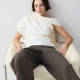 Woman sitting on a white chair wearing a white top and brown pants against a light gray background