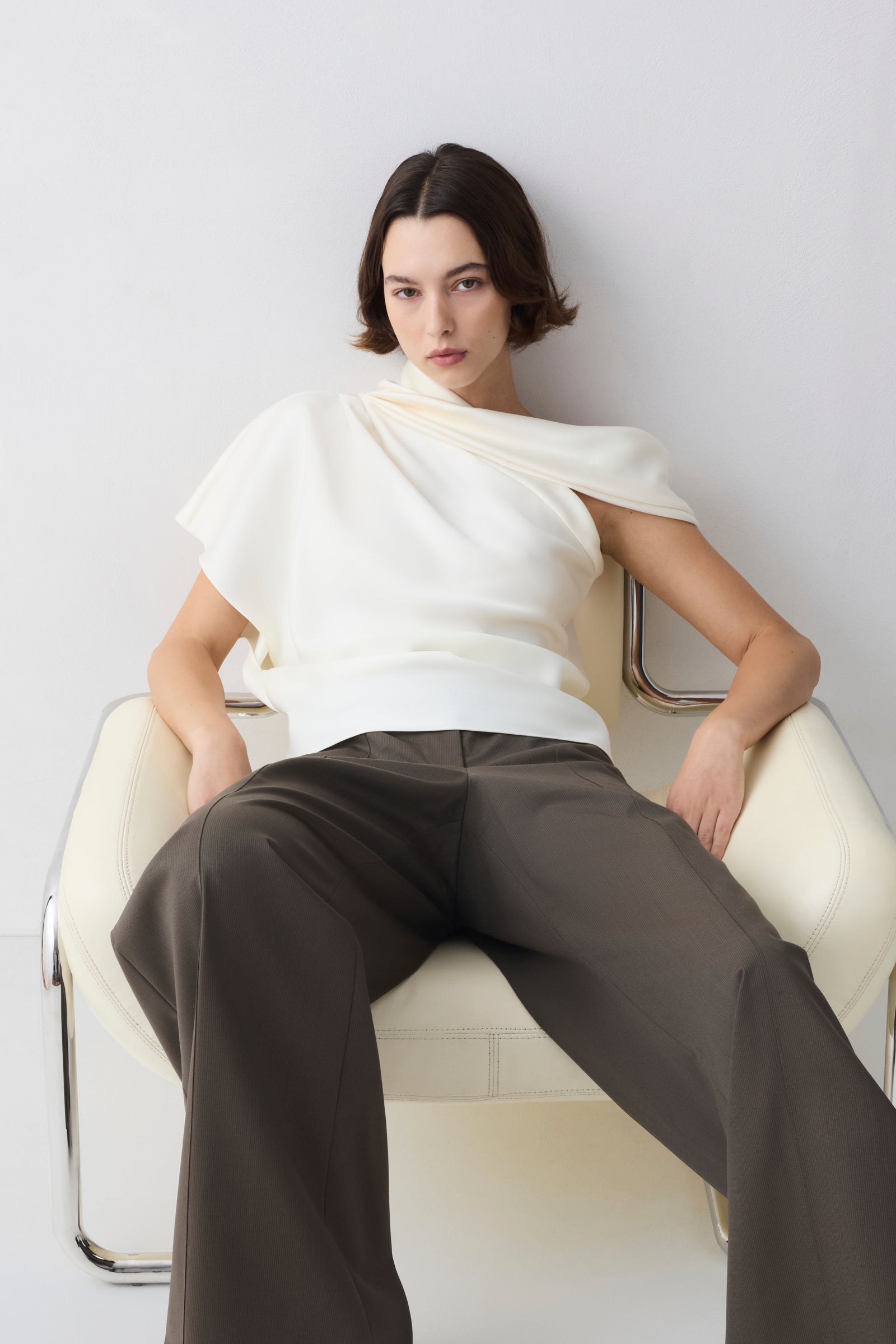 Woman sitting on a white chair wearing a white top and brown pants against a light gray background