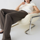 Woman sitting on a modern chair wearing a white top and brown pants against a white background