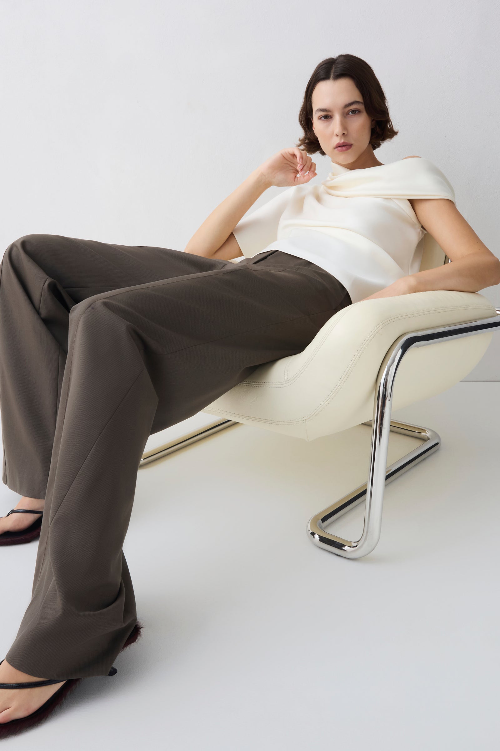 Woman sitting on a modern chair wearing a white top and brown pants against a white background