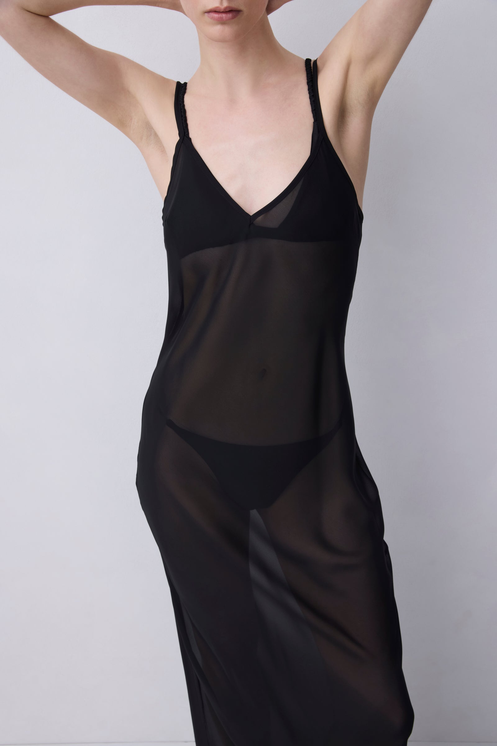 A model wearing a sheer black slip dress over black lingerie, photographed against a plain light gray studio background.
