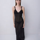 A model stands against a light gray studio background wearing a sheer black slip-style lingerie set with thin straps, a deep V-neckline, and a long flowing skirt.
