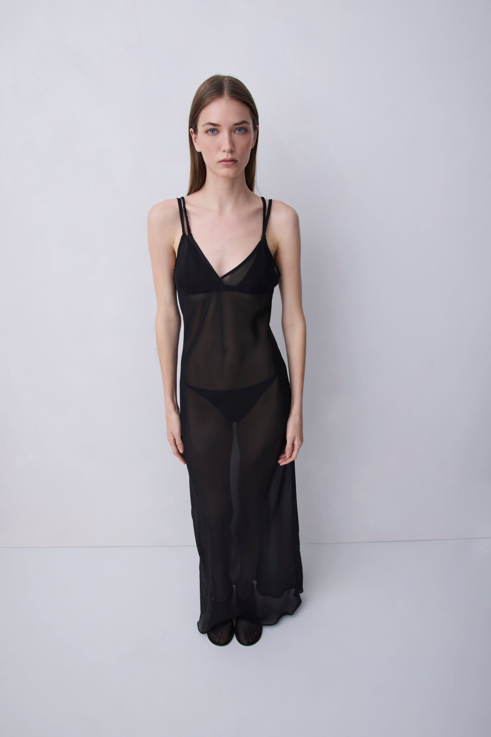 A model stands against a light gray studio background wearing a sheer black slip-style lingerie set with thin straps, a deep V-neckline, and a long flowing skirt.