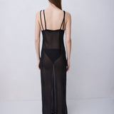 A woman stands with her back to the camera wearing a sheer black maxi slip dress with thin shoulder straps and a solid black panel at the waist, photographed against a plain light gray studio background.