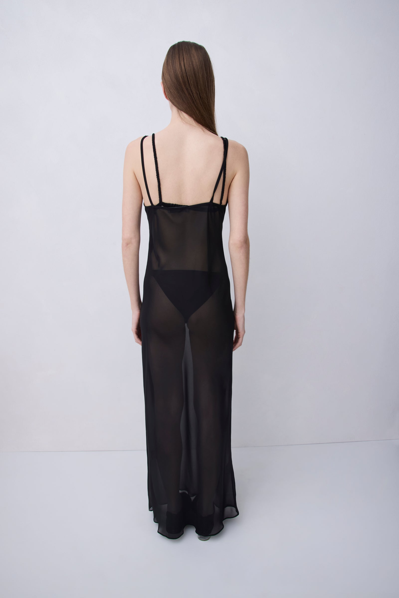 A woman stands with her back to the camera wearing a sheer black maxi slip dress with thin shoulder straps and a solid black panel at the waist, photographed against a plain light gray studio background.