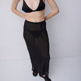 A model wearing a black sheer maxi skirt over a black bralette, standing against a light gray studio background.