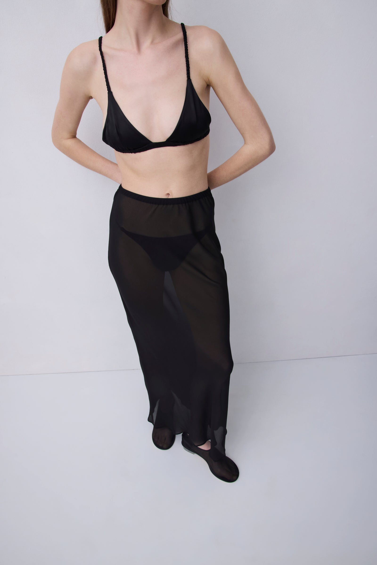 A model wearing a black sheer maxi skirt over a black bralette, standing against a light gray studio background.