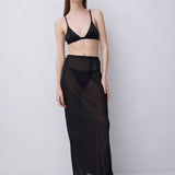 A model stands against a light gray studio background wearing a black sheer maxi skirt with a wide waistband, paired with a black bralette and black slip-on shoes.