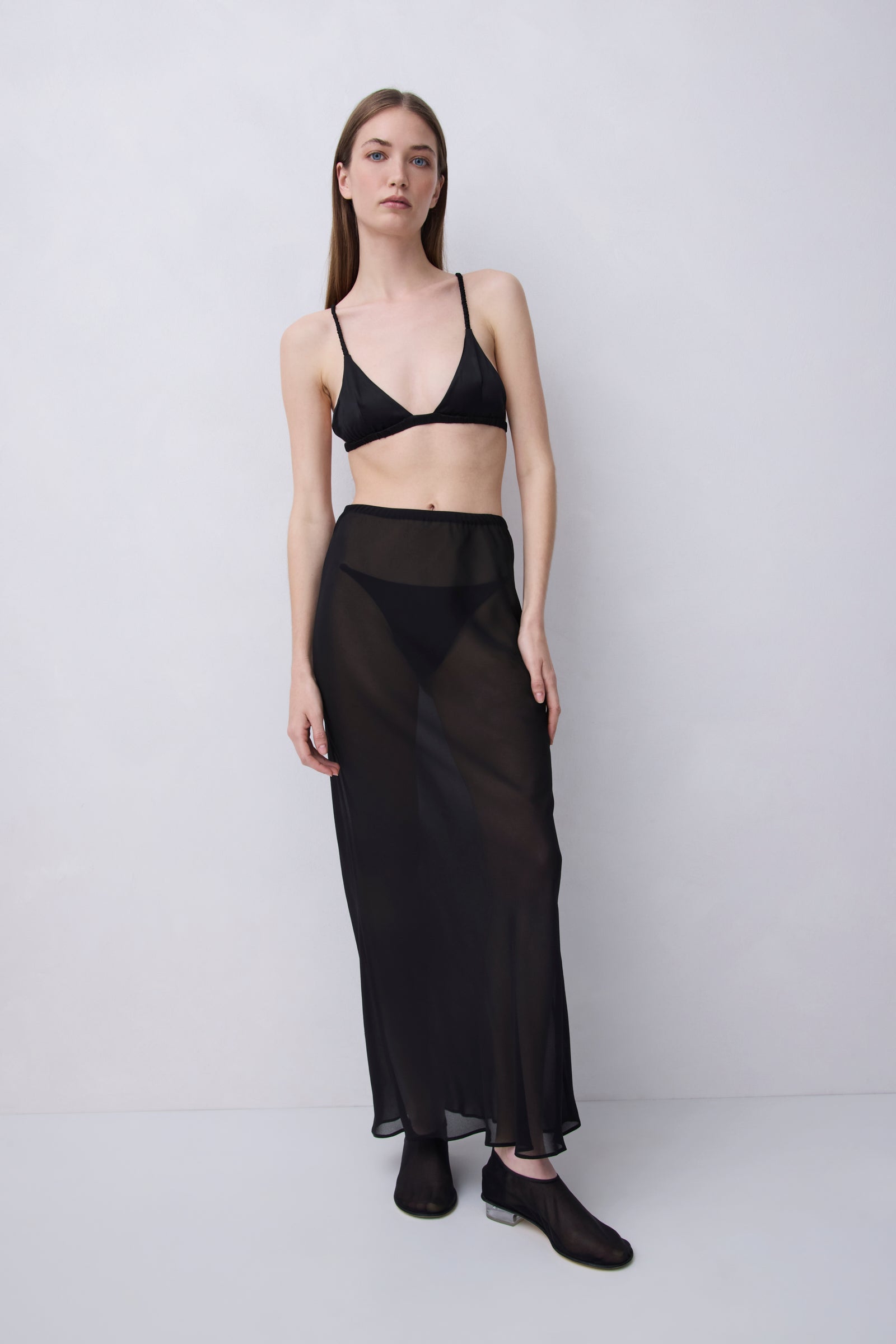 A model stands against a light gray studio background wearing a black sheer maxi skirt with a wide waistband, paired with a black bralette and black slip-on shoes.