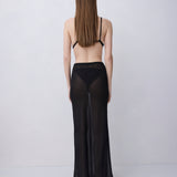 A woman viewed from behind wearing a sheer black maxi skirt with wide legs and a black bralette, standing against a plain light gray studio background.