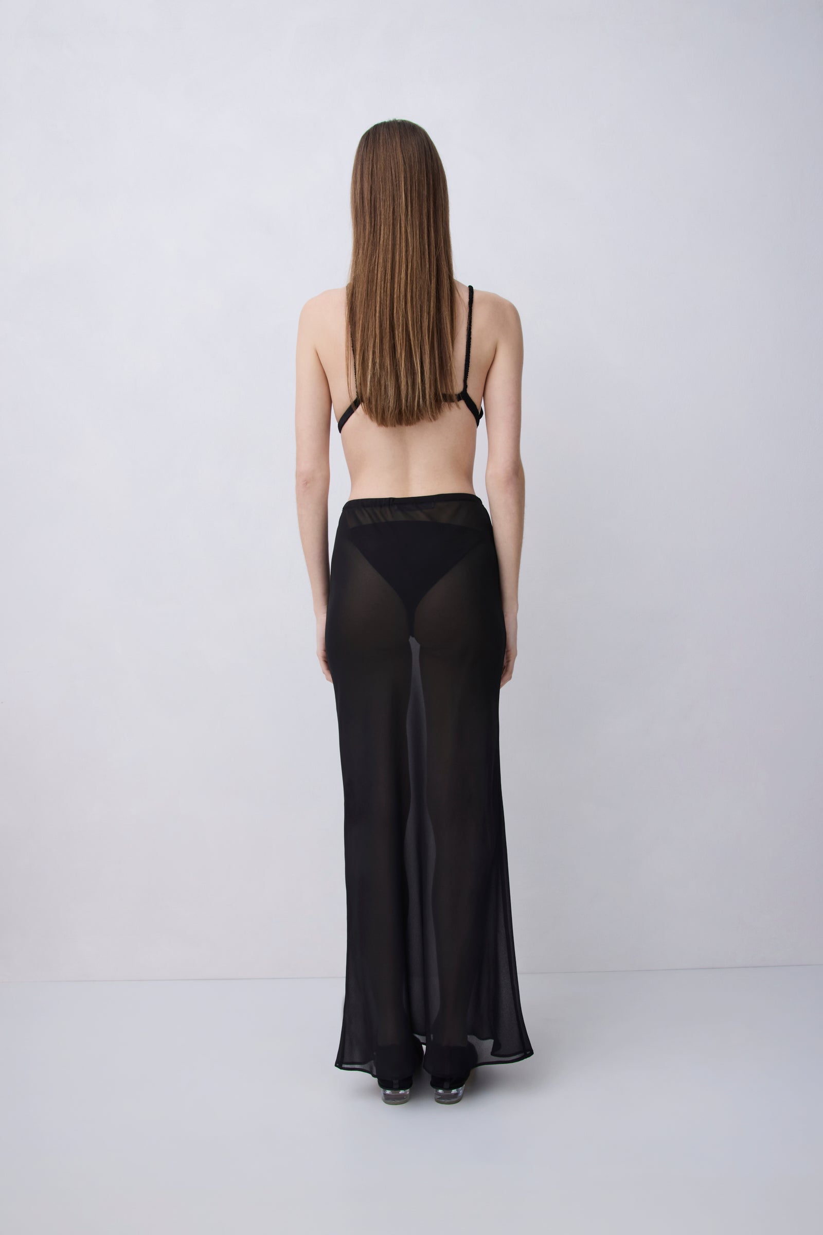 A woman viewed from behind wearing a sheer black maxi skirt with wide legs and a black bralette, standing against a plain light gray studio background.