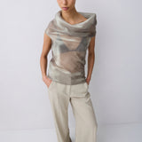 A model wearing a draped, semi-sheer metallic beige top paired with wide-leg beige trousers, standing against a plain light gray studio background.