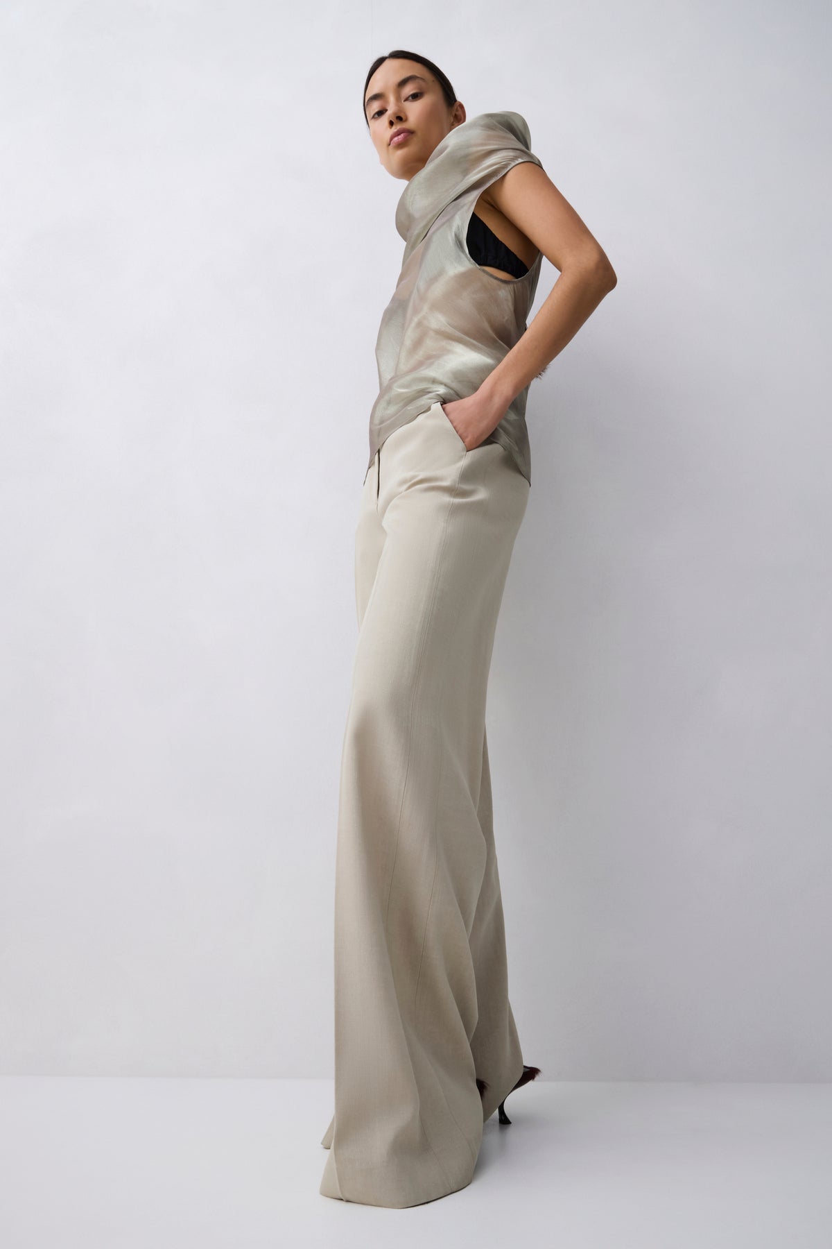 A model wearing a beige satin-like draped top and matching wide-leg trousers, posing against a light gray studio background.