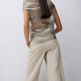 A model shown from behind wearing a metallic champagne-beige short-sleeve top and light beige wide-leg trousers against a light gray studio background.