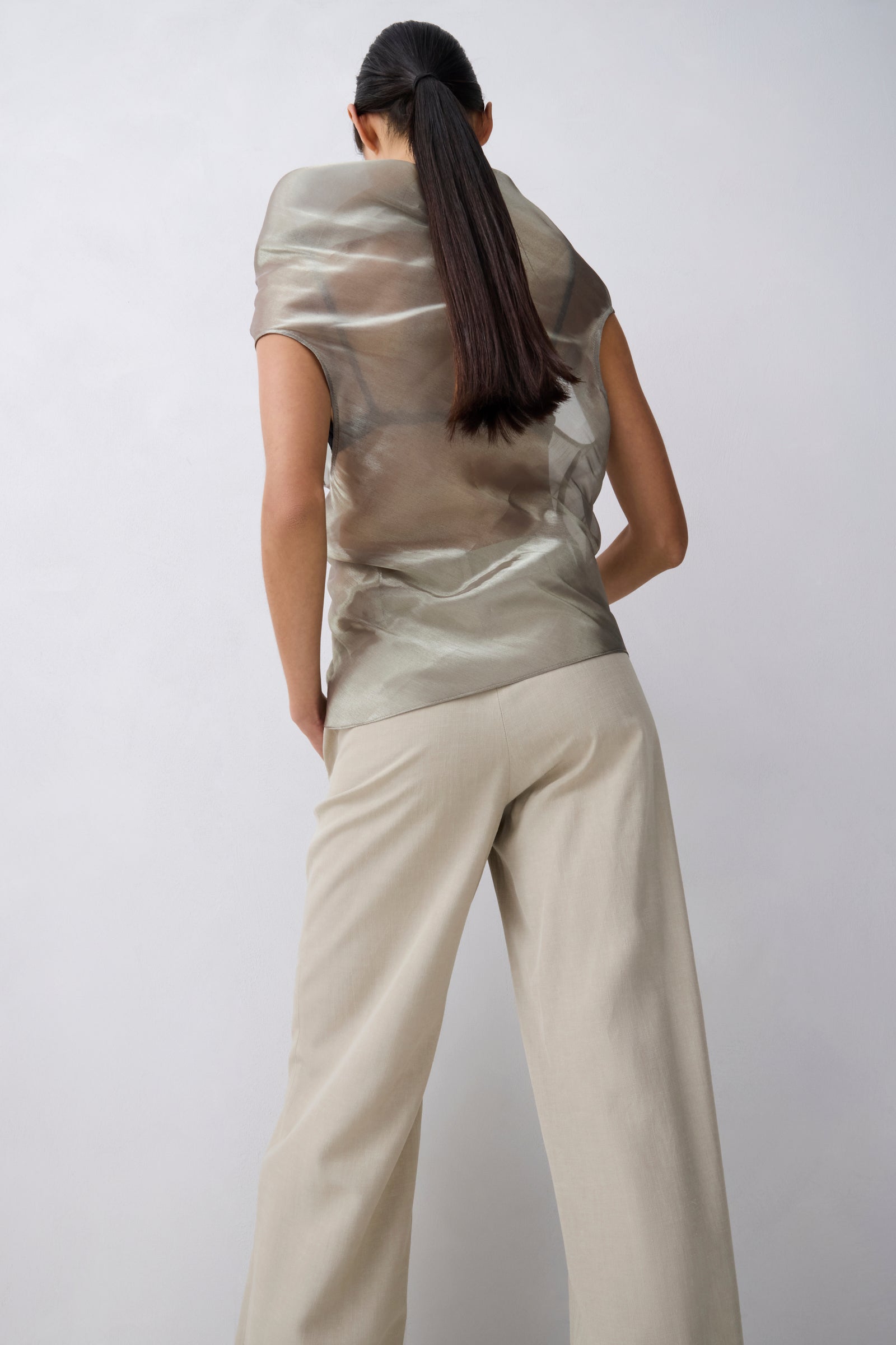 A model shown from behind wearing a metallic champagne-beige short-sleeve top and light beige wide-leg trousers against a light gray studio background.