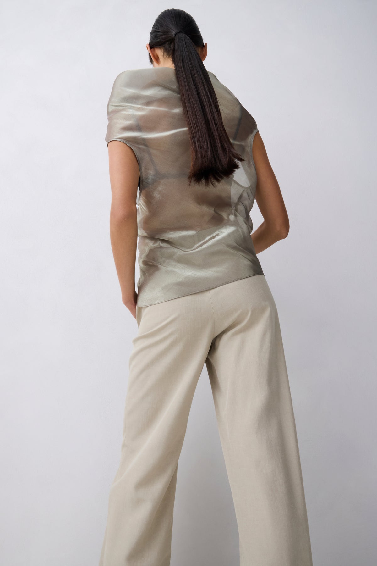 A model shown from behind wearing a metallic champagne-beige short-sleeve top and light beige wide-leg trousers against a light gray studio background.
