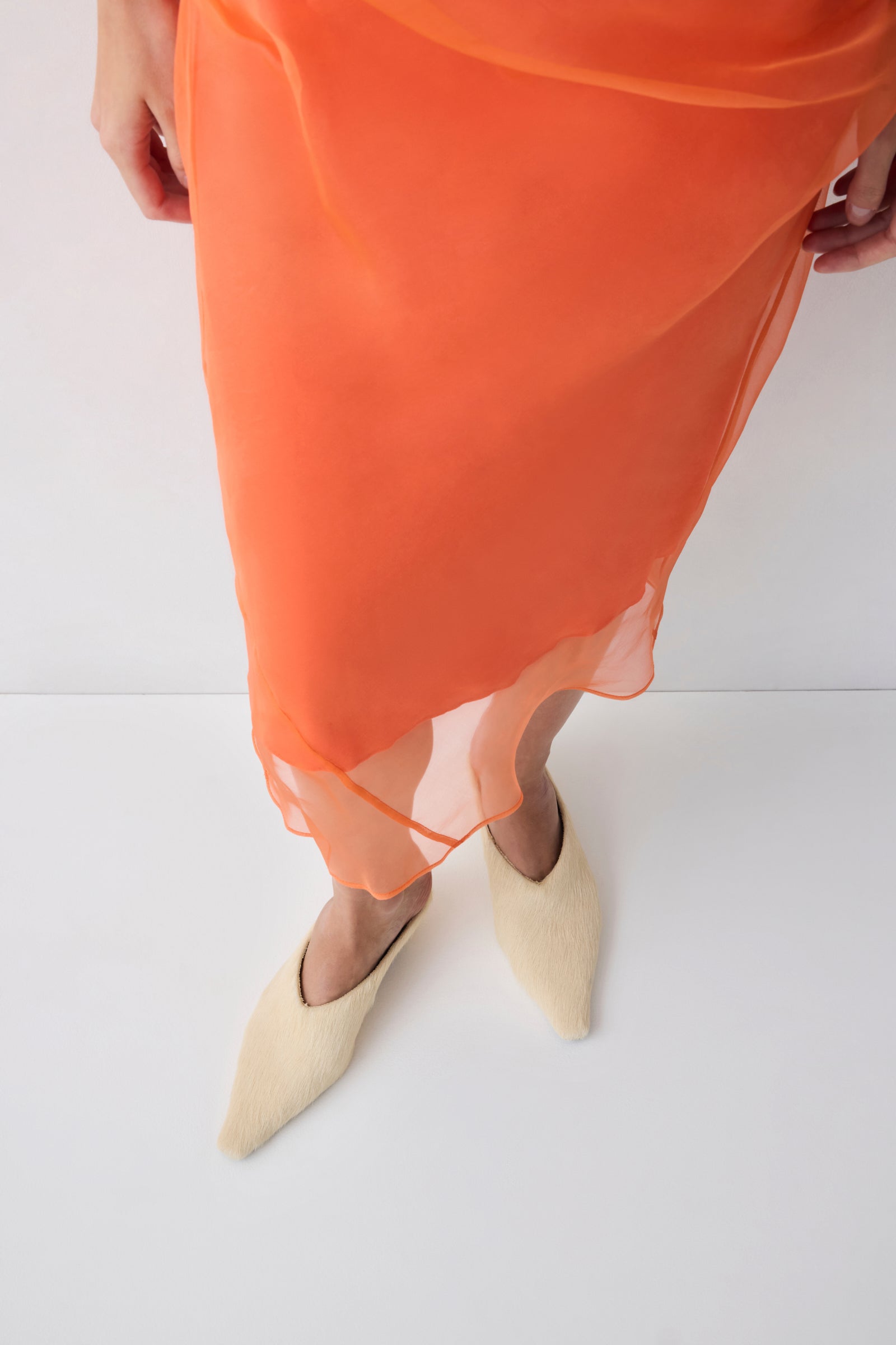 Person wearing an orange sheer skirt with beige pointed-toe shoes on a white background