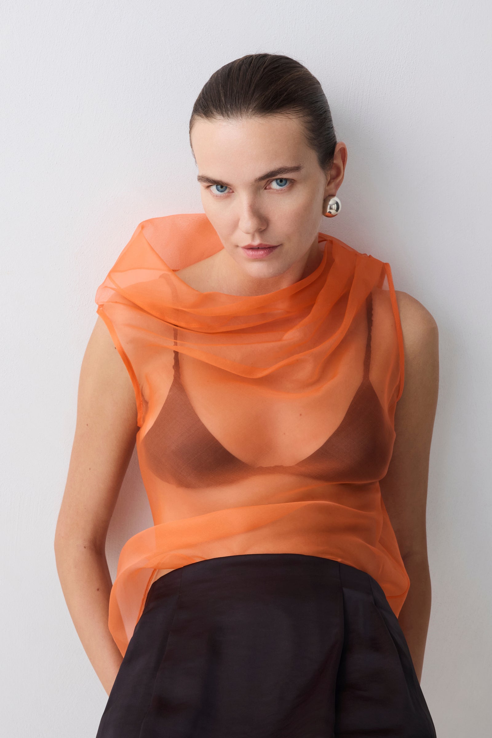 Woman wearing an orange sheer top and black pants against a white background