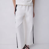 White wide-leg trousers with black side stripes, worn by a model with a white top and black strappy heels against a white studio background.