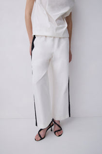 White wide-leg trousers with black side stripes, worn by a model with a white top and black strappy heels against a white studio background.