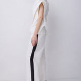 A model is shown in profile wearing a white sleeveless top and matching wide-leg trousers with a bold black vertical stripe down the side, paired with black strappy high-heeled sandals, set against a plain light gray studio background.