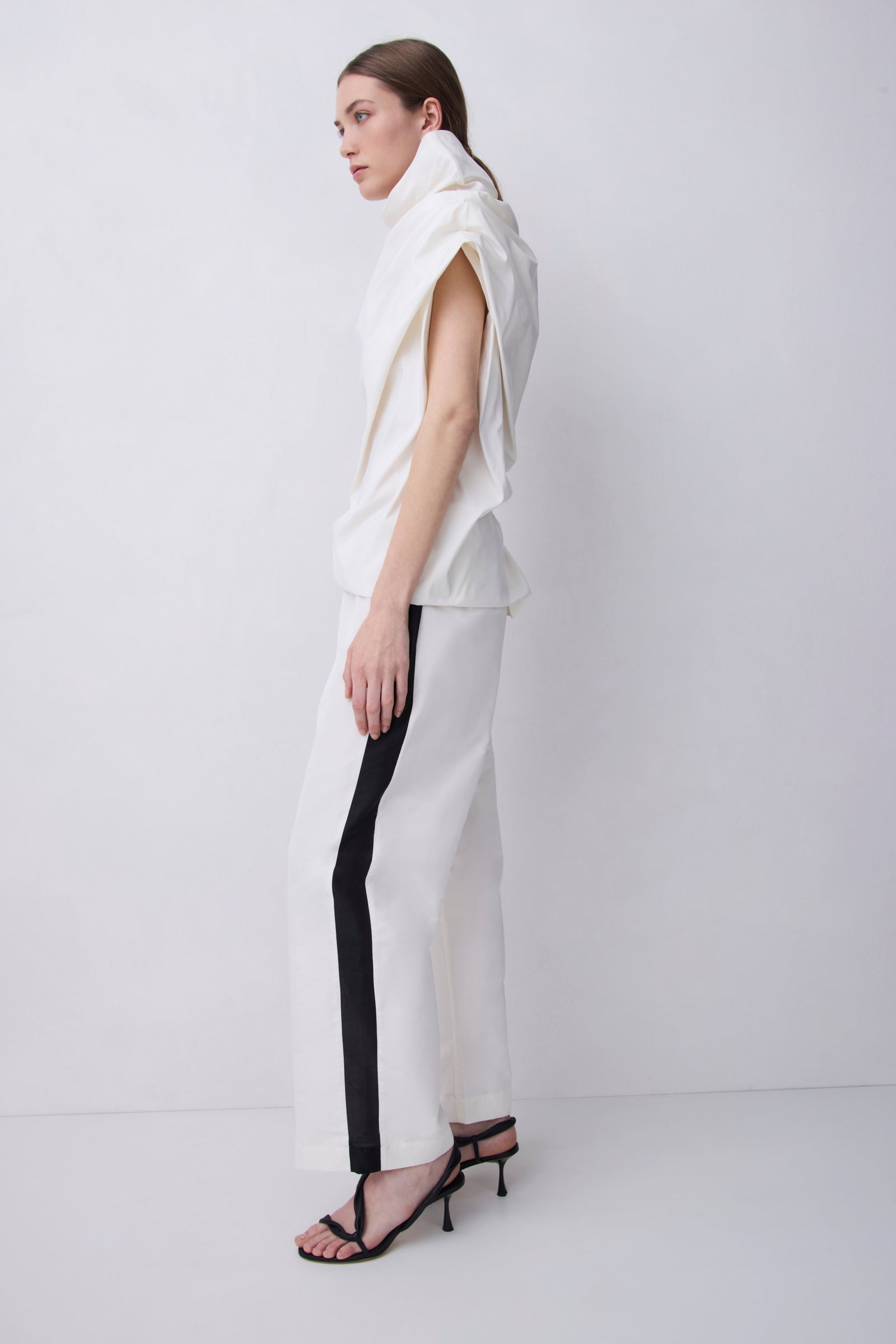 A model is shown in profile wearing a white sleeveless top and matching wide-leg trousers with a bold black vertical stripe down the side, paired with black strappy high-heeled sandals, set against a plain light gray studio background.