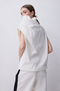 A model shown from the back wearing a sleeveless, high-waisted white top with a dramatic draped collar and matching white trousers with a black side stripe, set against a plain light gray studio background.