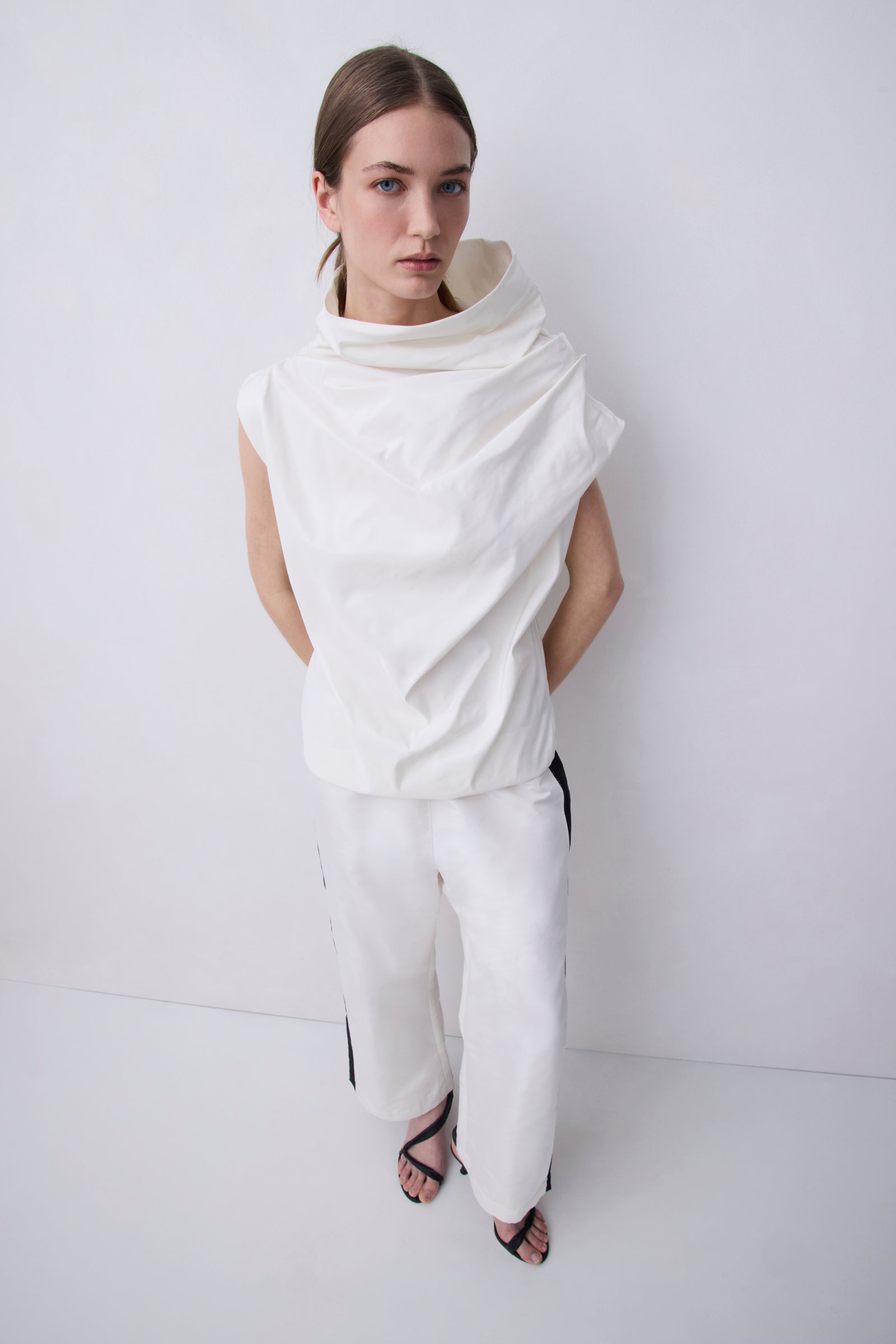 A model wearing a white draped sleeveless top and matching wide-leg trousers with black side stripes, standing against a plain white studio background.