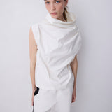 A model wearing a white sleeveless draped top with a high draped collar and matching white draped trousers, standing against a light gray studio background.