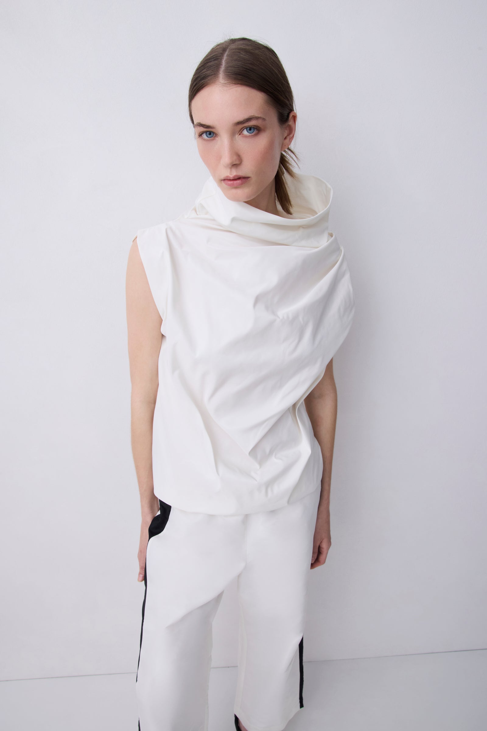 A model wearing a white sleeveless draped top with a high draped collar and matching white draped trousers, standing against a light gray studio background.