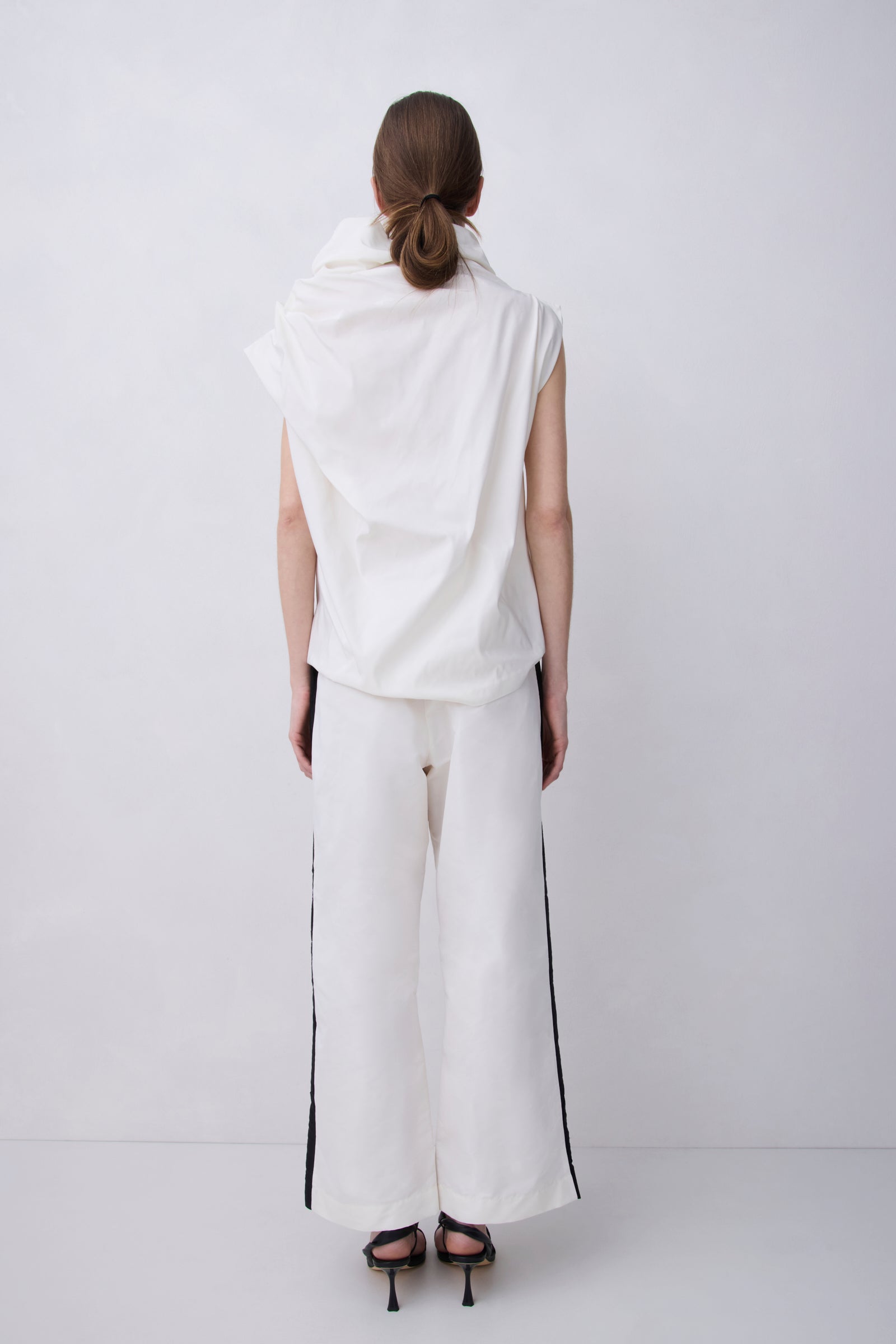 A model viewed from the back wearing a white sleeveless draped top and matching white wide-leg trousers with black side stripes, paired with black high-heeled sandals, against a plain light gray studio background.