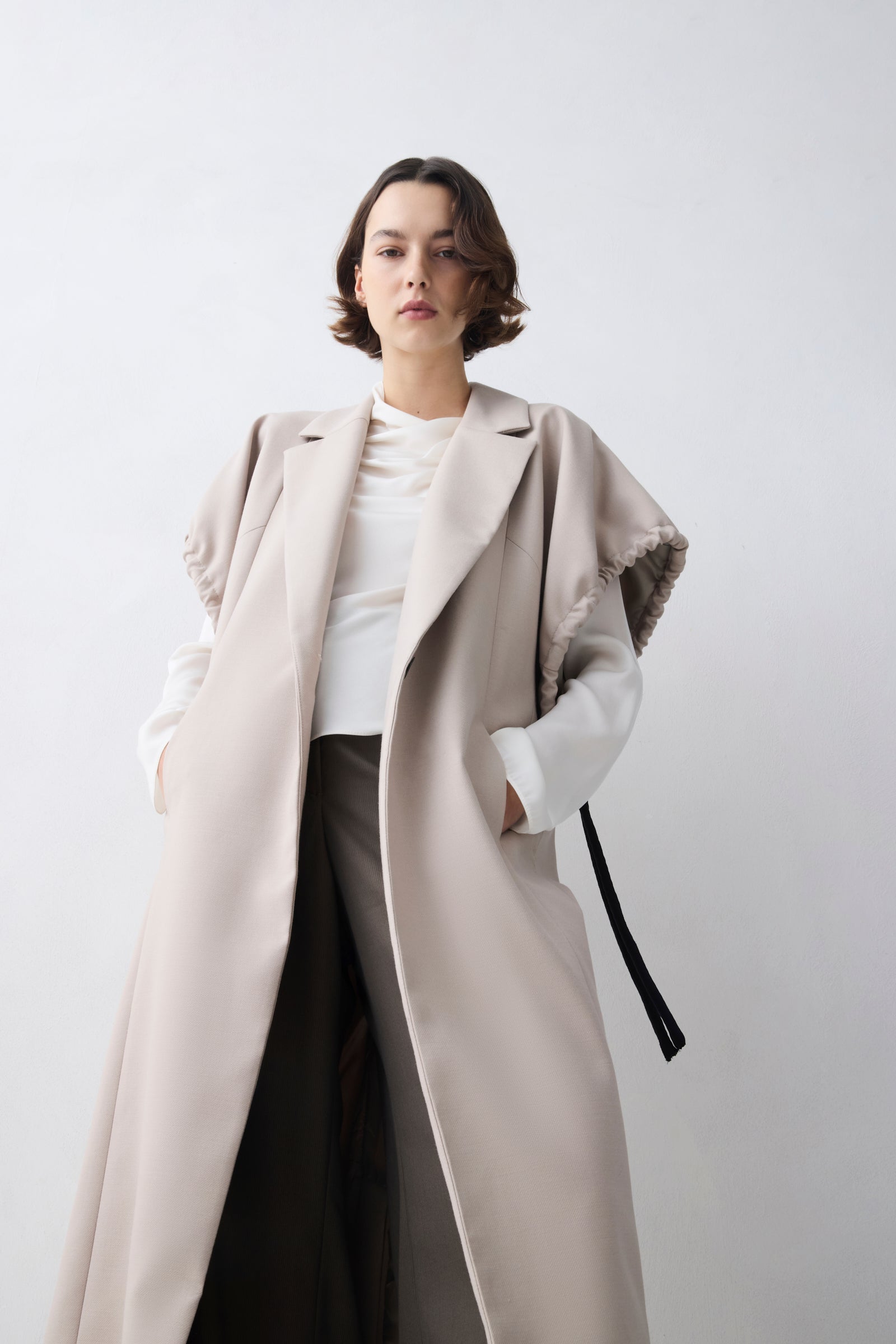 Person wearing a long beige coat over a white top and dark pants on a light gray background
