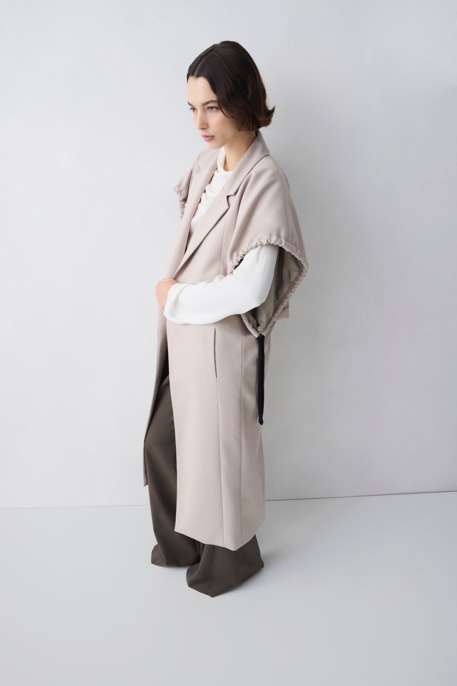 Person wearing a beige coat and pants with a white shirt against a plain background