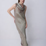 A woman modeling a sleeveless, floor-length draped dress in a shimmering taupe-beige color, standing against a plain light gray studio background.