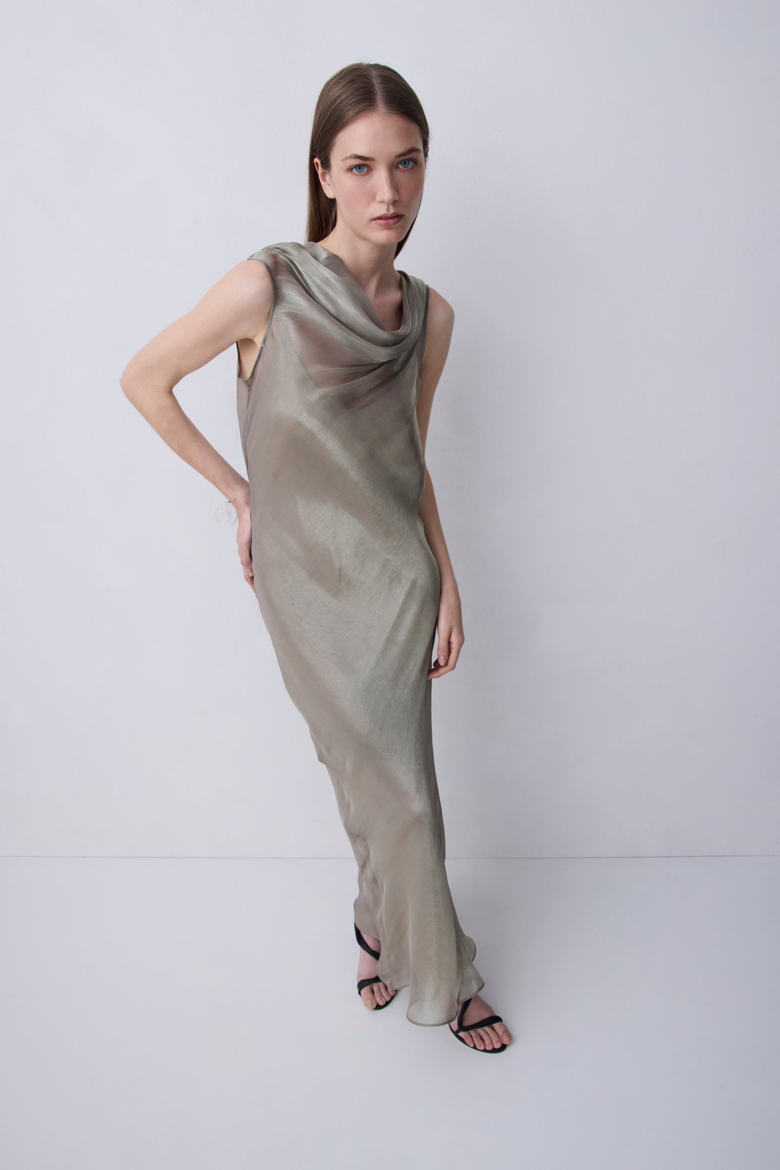 A woman modeling a sleeveless, floor-length draped dress in a shimmering taupe-beige color, standing against a plain light gray studio background.