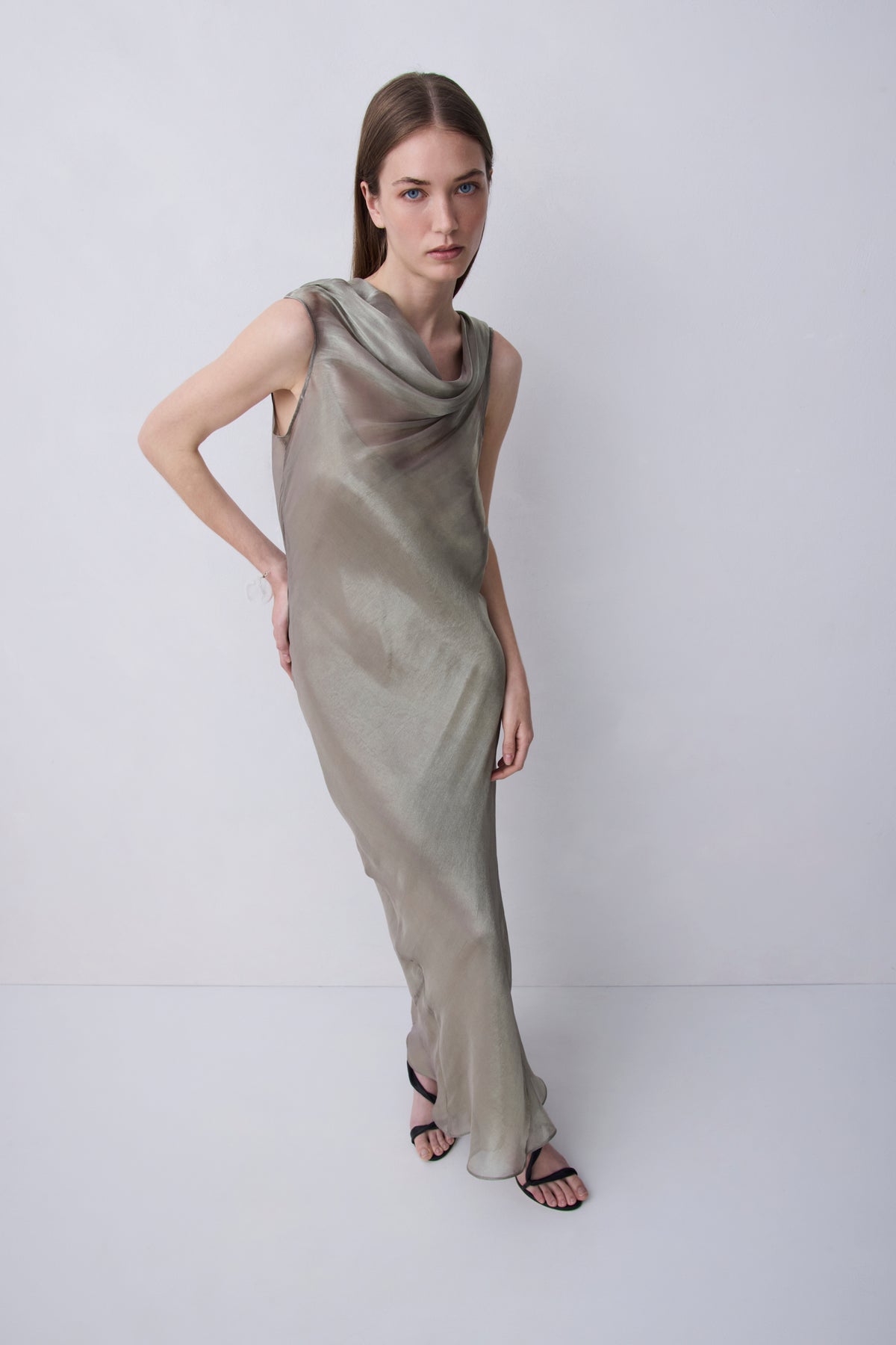 A woman modeling a sleeveless, floor-length draped dress in a shimmering taupe-beige color, standing against a plain light gray studio background.