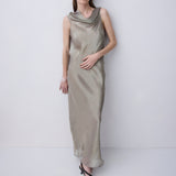 A woman modeling a sleeveless, cowl-neck maxi dress in a shimmering champagne-gold satin, standing against a plain light gray studio background.